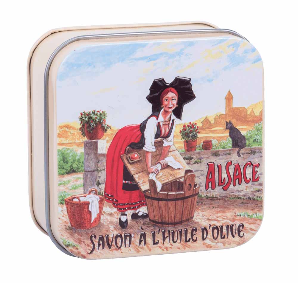 La Savonnerie de Nyons presents its exquisite cotton flower soap, encased in a charming vintage tin titled 'L'Alsacienne'. This tin, adorned with Luc Mazan's artwork, showcases a traditional Alsatian woman performing the timeless task of washing clothes, capturing the essence of Alsace's rich cultural heritage.